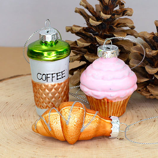 Drei Glas Weihnachtsanhänger mit Kaffee, Croissant und rosa Cupcake auf einer Holzscheibe