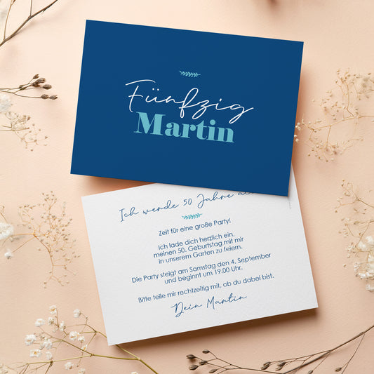 Personalisierte Einladungskarte zum 50. Geburtstag mit stilvollem Design in Dunkelblau. Die Vorderseite zeigt den Schriftzug „Fünfzig Martin“ in einer eleganten Schriftart. Die Rückseite bietet Platz für individuellen Einladungstext und Details zur Feier. Die Karte liegt auf einer beigen Oberfläche mit dekorativen Trockenblumen, passend zur festlichen Atmosphäre.
