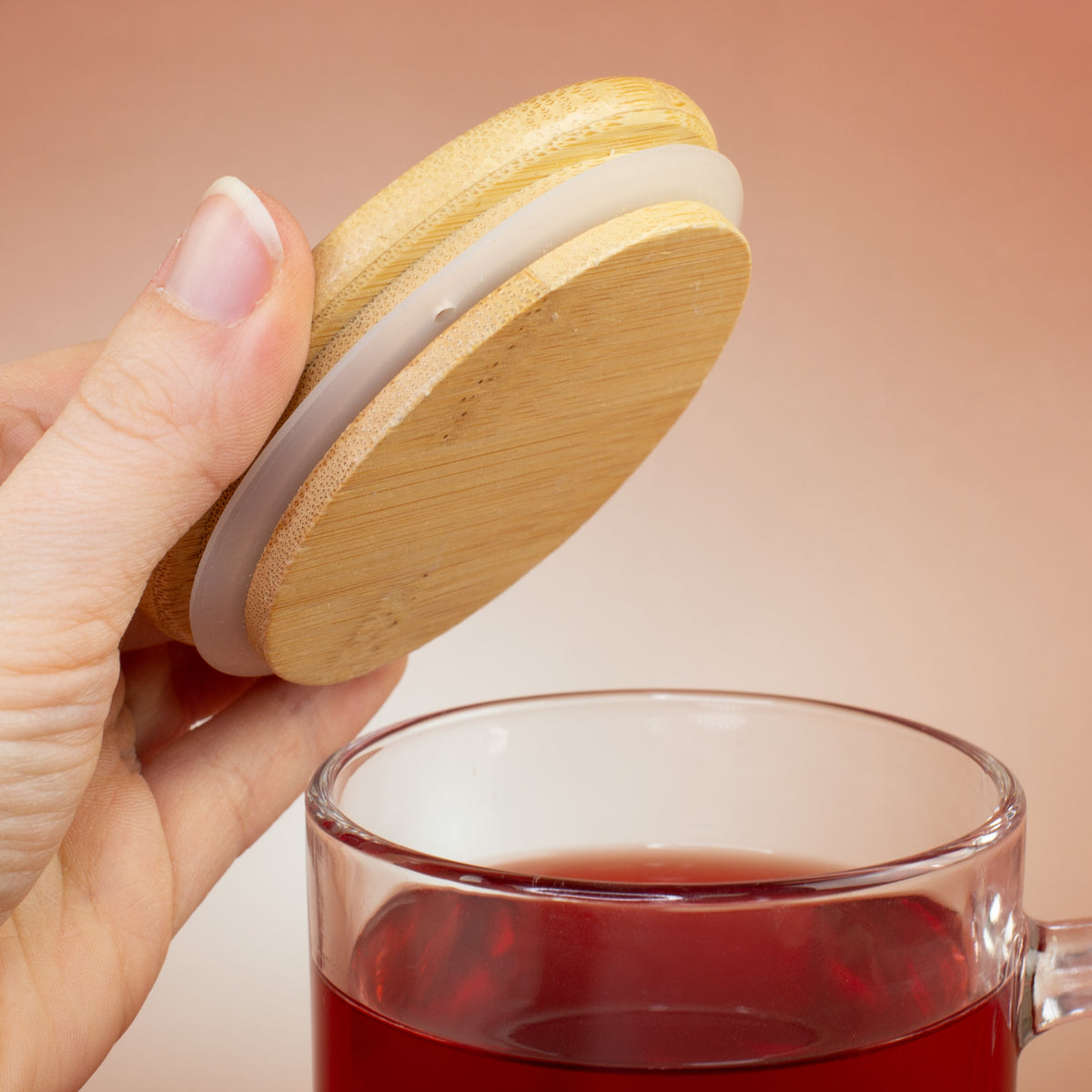 Hand hält runden Bambusdeckel mit Dichtungsring über einer Glastasse mit rotem Tee