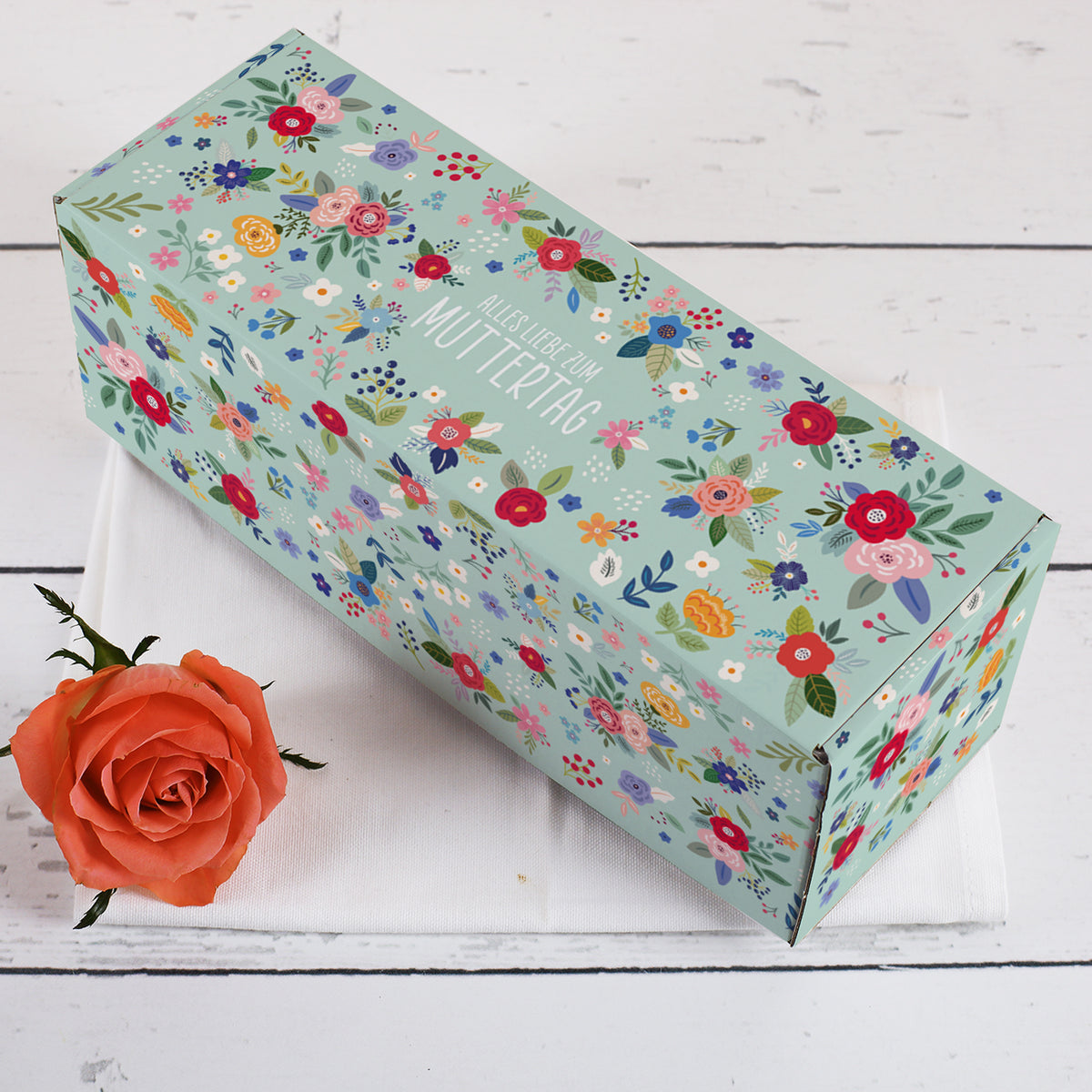 Längliche Geschenkbox zum Muttertag mit floraler Bedruckung und Aufschrift Alles Liebe zum Muttertag – Dekoszene mit orangener Rose