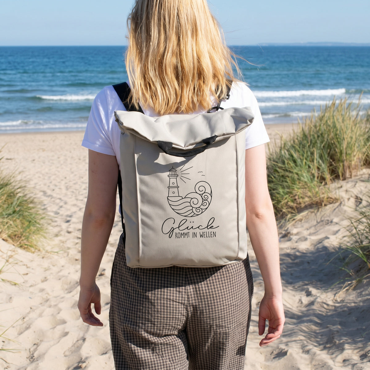 Beiger Rucksack mit Leuchtturm Motiv wird am Strand getragen und eignet sich für Urlaub, Wandern und Freizeit