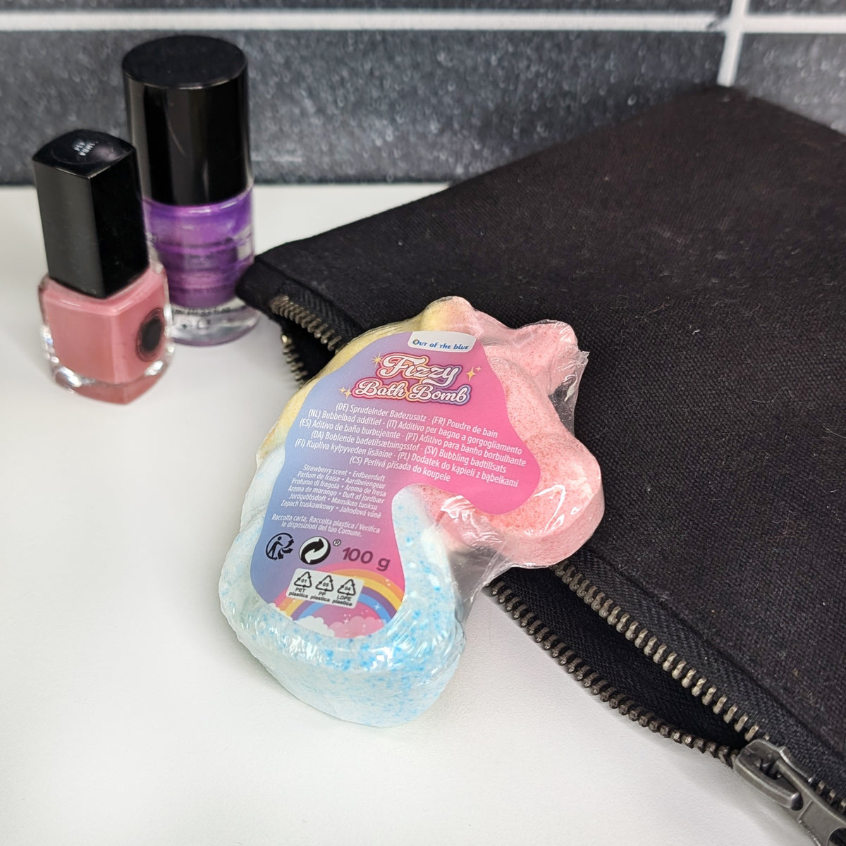 Einhorn-Badekugel in Verpackung mit Etikett, arrangiert mit Nagellack und schwarzem Täschchen