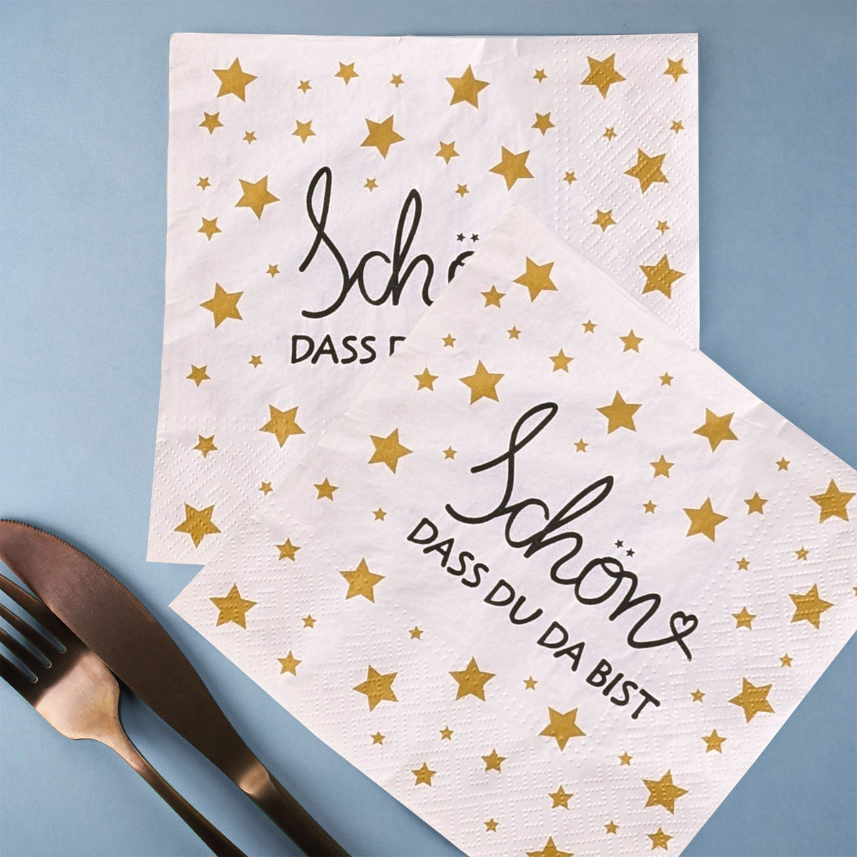 Weiße Papierservietten mit goldenen Sternen und dem Aufdruck ‚Schön, dass du da bist‘, dekorativ auf blauem Untergrund mit Besteck arrangiert.