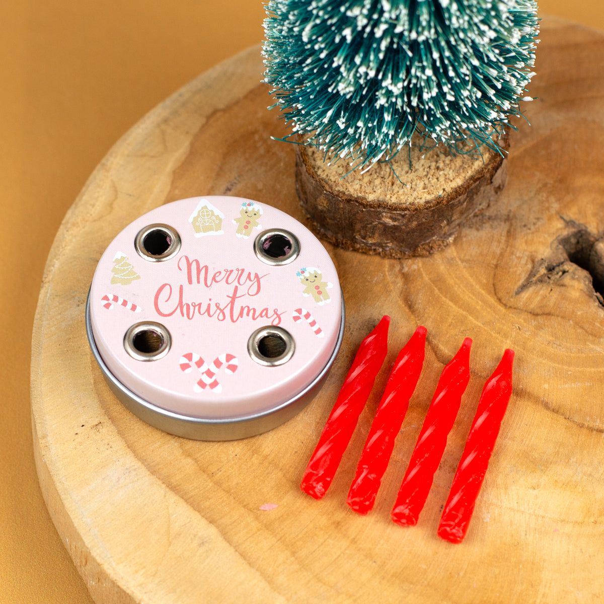 Mini-Adventskranz mit rosafarbener „Merry Christmas“-Aufschrift, umgeben von Weihnachtsmotiven, liegend auf einem Holzbrett mit vier roten Kerzen daneben.