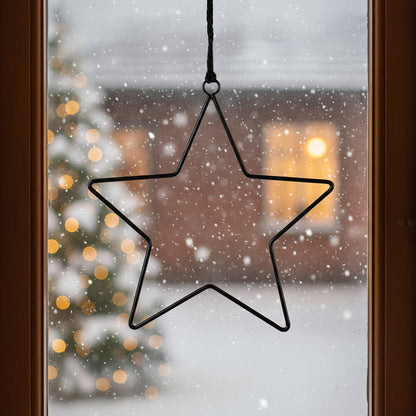 Ein schwarzer Metallstern hängt mit einer Schnur im Fenster. Draußen ist eine verschneite Winterlandschaft mit Weihnachtsbaum und Lichtern zu sehen.