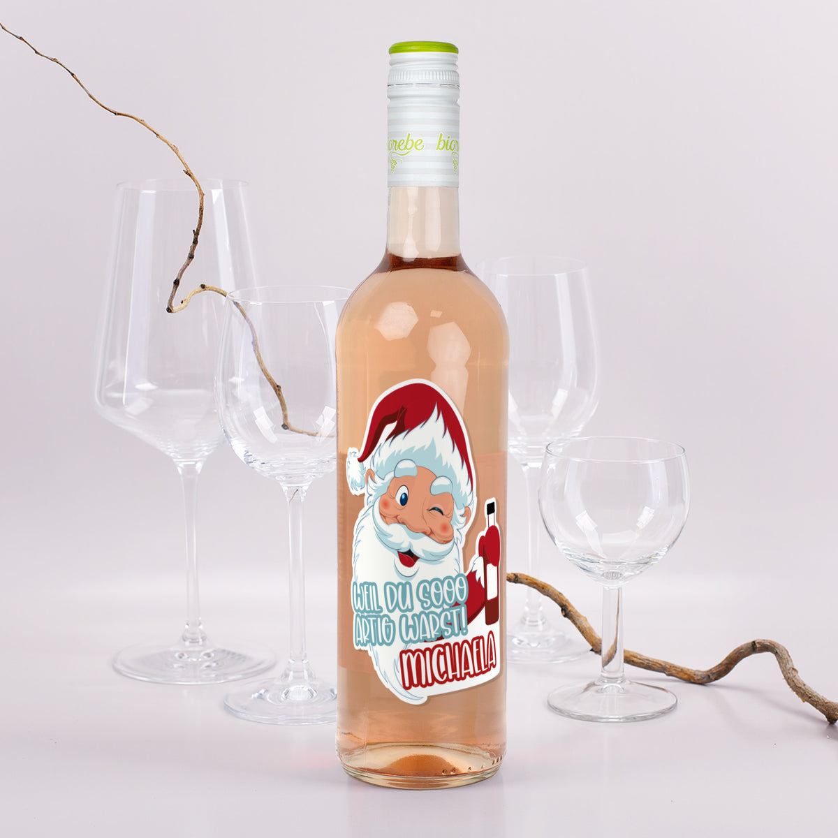 Roséweinflasche mit Weihnachtsmann-Etikett „Weil du sooo artig warst! Michaela“, dekoriert mit Gläsern und Zweigen.