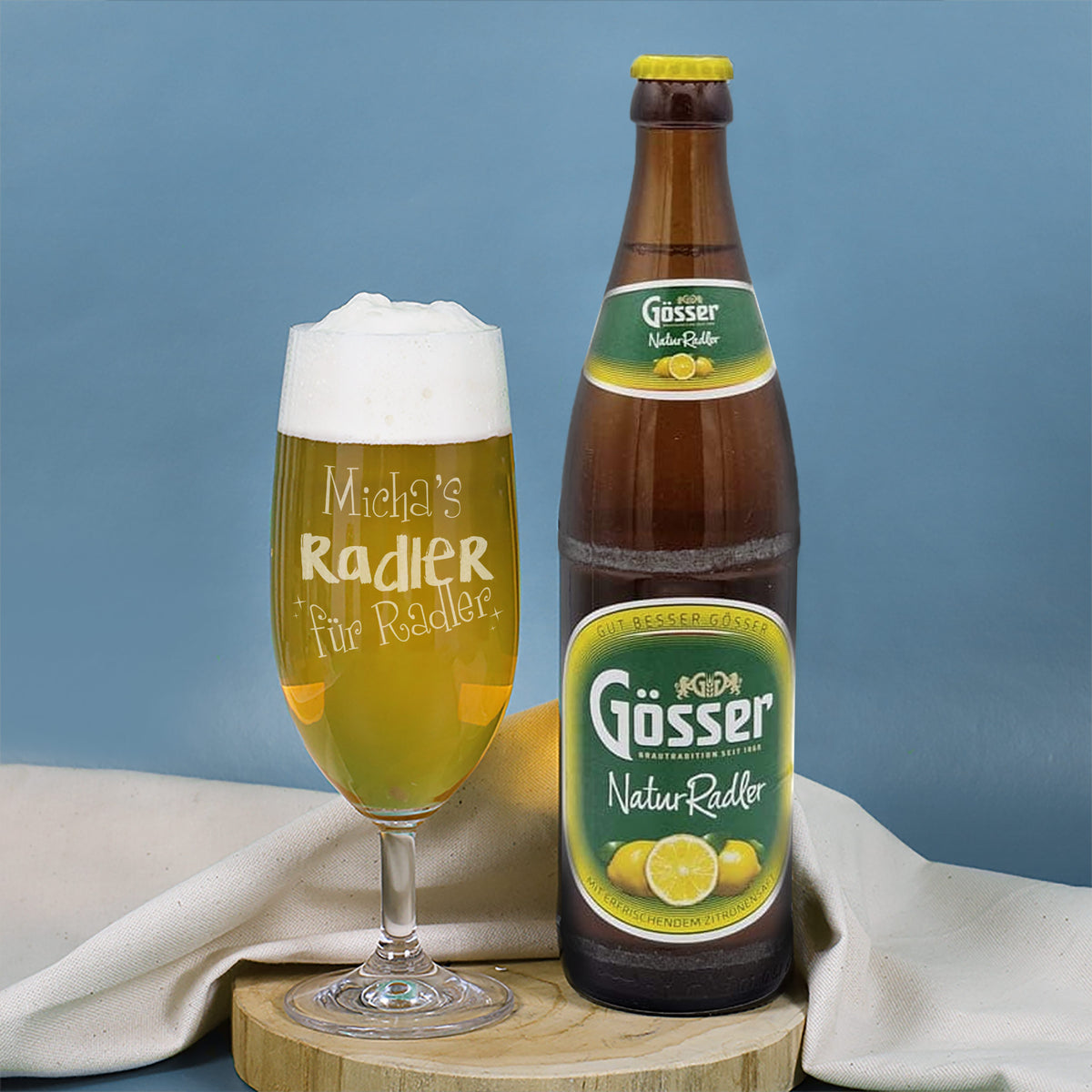 Bierglas „Radler für Radler“ mit Gravur und Flasche Gösser NaturRadler auf Holzbrett vor blauem Hintergrund