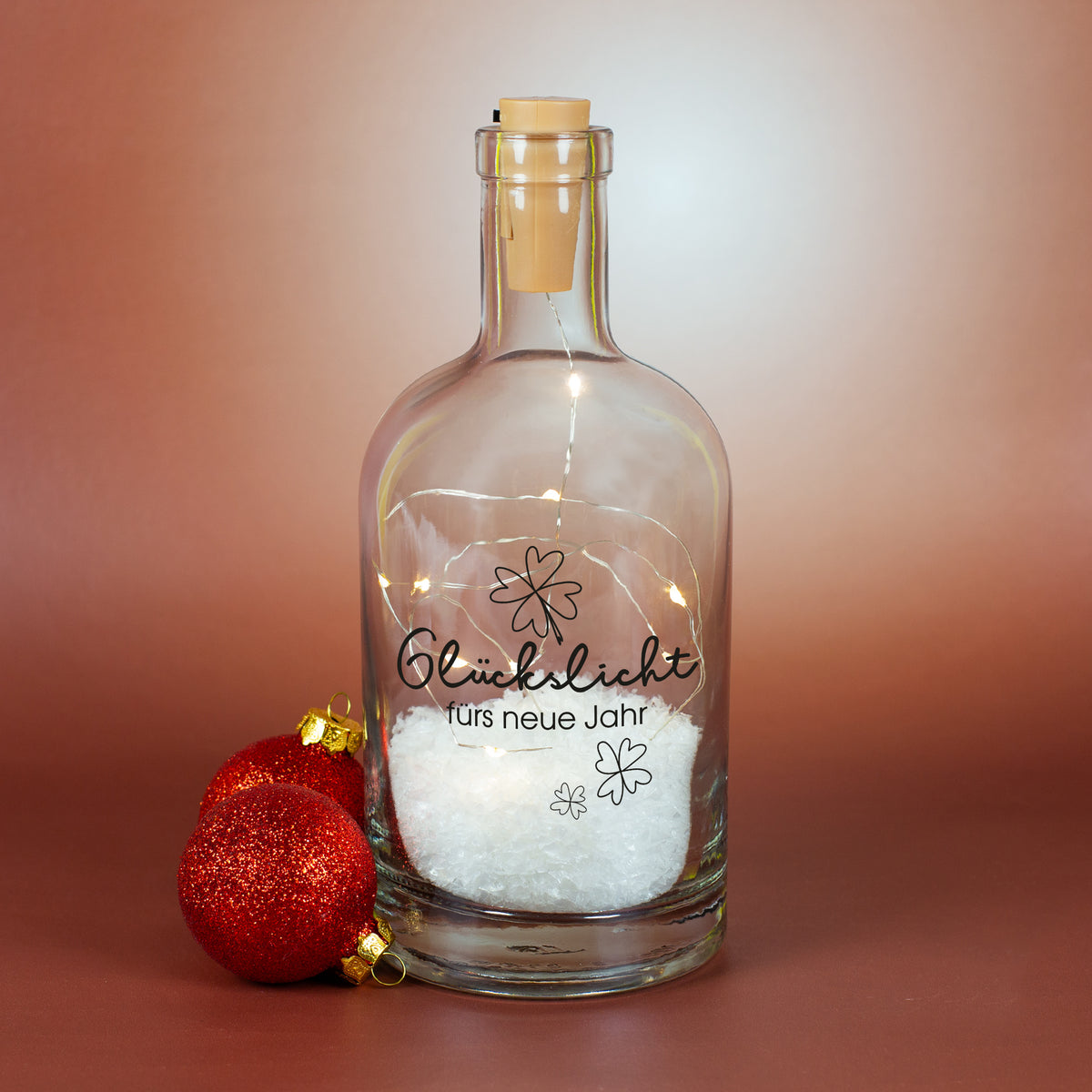 Personalisierte LED-Glückslicht-Karaffe mit Kunstschnee und leuchtender Lichterkette, Aufdruck „Glückslicht fürs neue Jahr“, fotografiert vor warmem Hintergrund und roten Weihnachtskugeln.