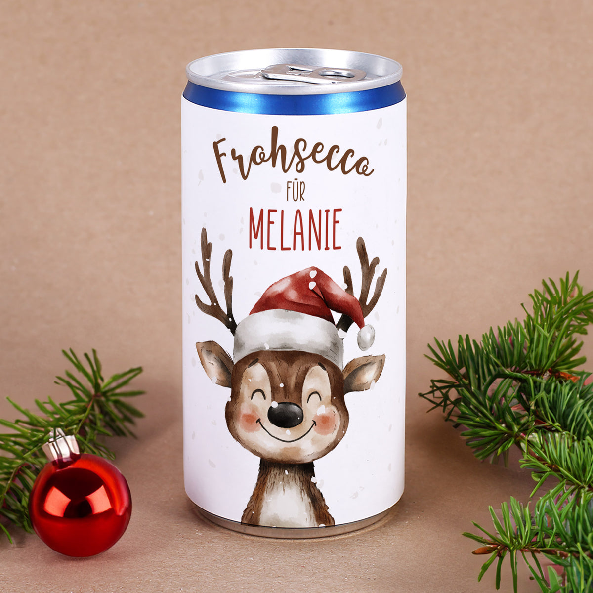 Weihnachtliche Prosecco-Dose mit Rentier-Illustration und Schriftzug „Frohsecco“ auf braunem Hintergrund mit Tannenzweig und Christbaumkugel