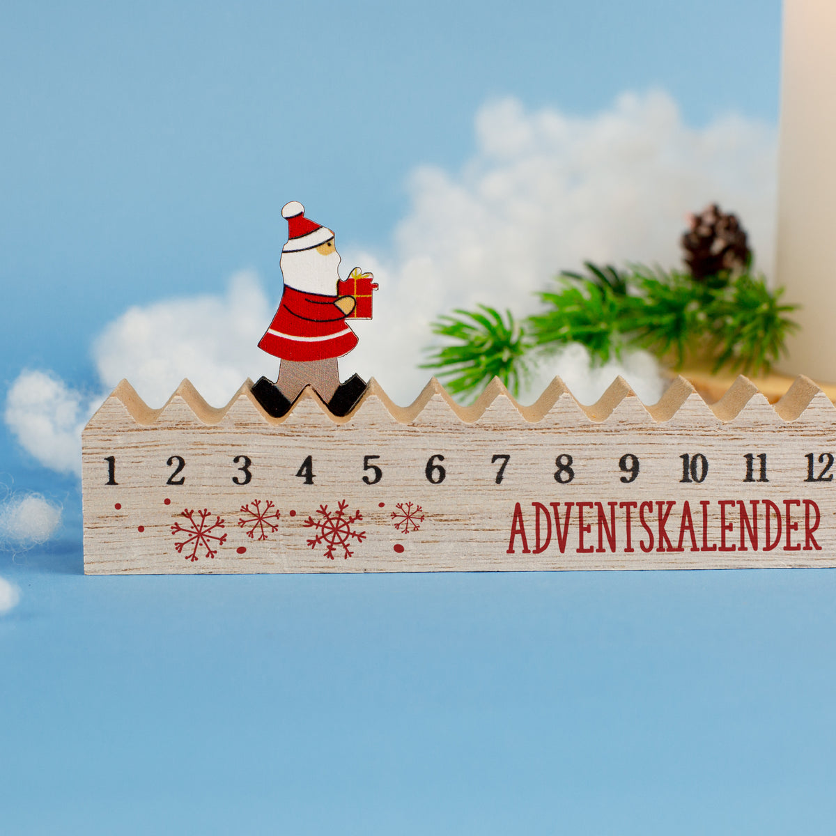 Adventskalender aus Holz mit Zahlenreihe und Santa-Figur, dekoriert mit grünen Zweigen und Schneewatte.