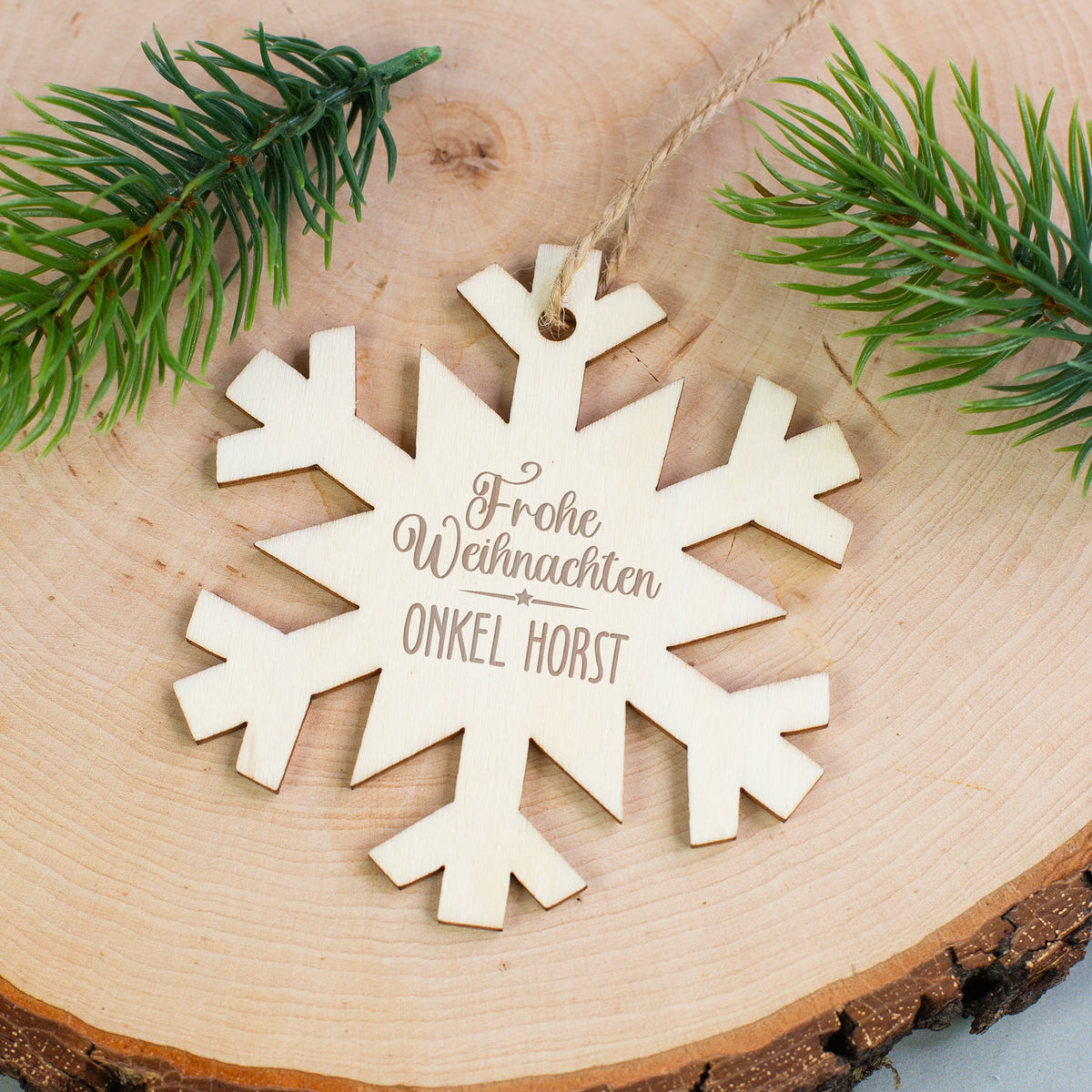 Personalisierter Holzanhänger Schneeflocke mit Gravur „Frohe Weihnachten Onkel Horst“, liegend auf Holzscheibe mit Tannenzweigen.