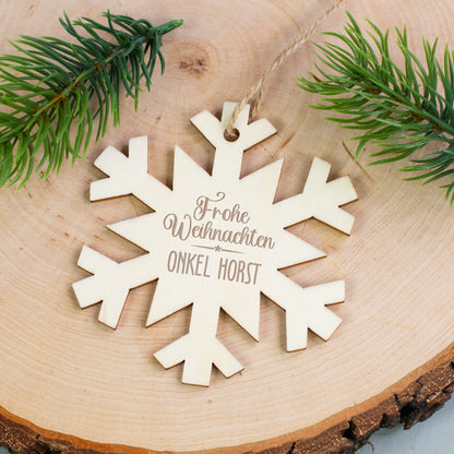 Personalisierter Holzanhänger Schneeflocke mit Gravur „Frohe Weihnachten Onkel Horst“, liegend auf Holzscheibe mit Tannenzweigen.