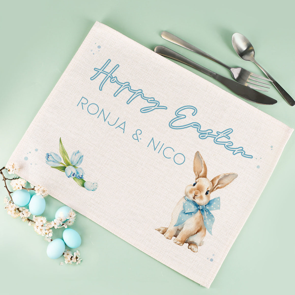 Personalisiertes Jute Platzdeckchen zu Ostern mit Hasenmotiv, blauer Schleife, Blumenillustration und individuellem Wunschtext auf mintgrünem Hintergrund