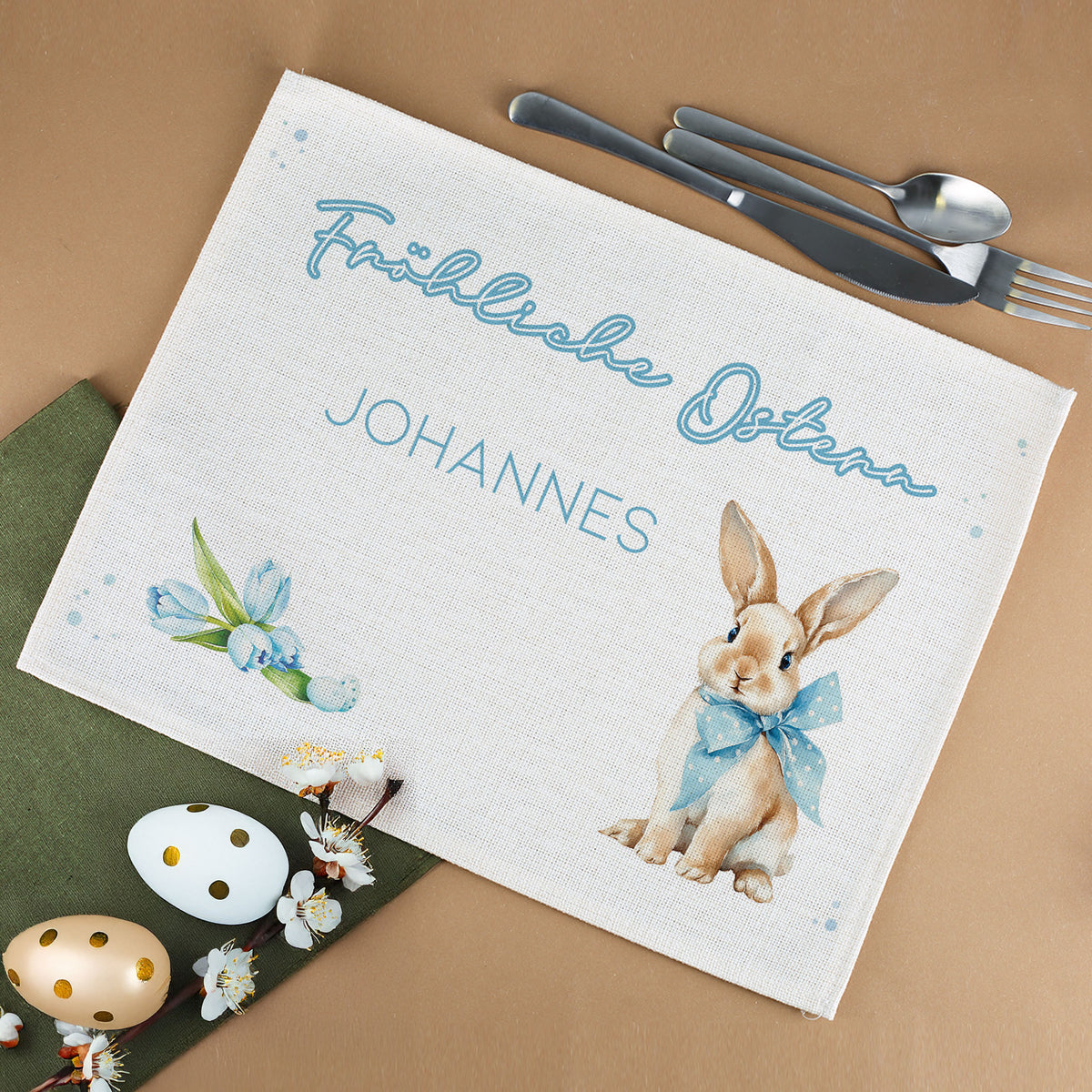 Personalisiertes Oster Platzdeckchen aus Jute mit Wunschtext, Hasenmotiv und Blumenillustration neben Ostereiern und Besteck
