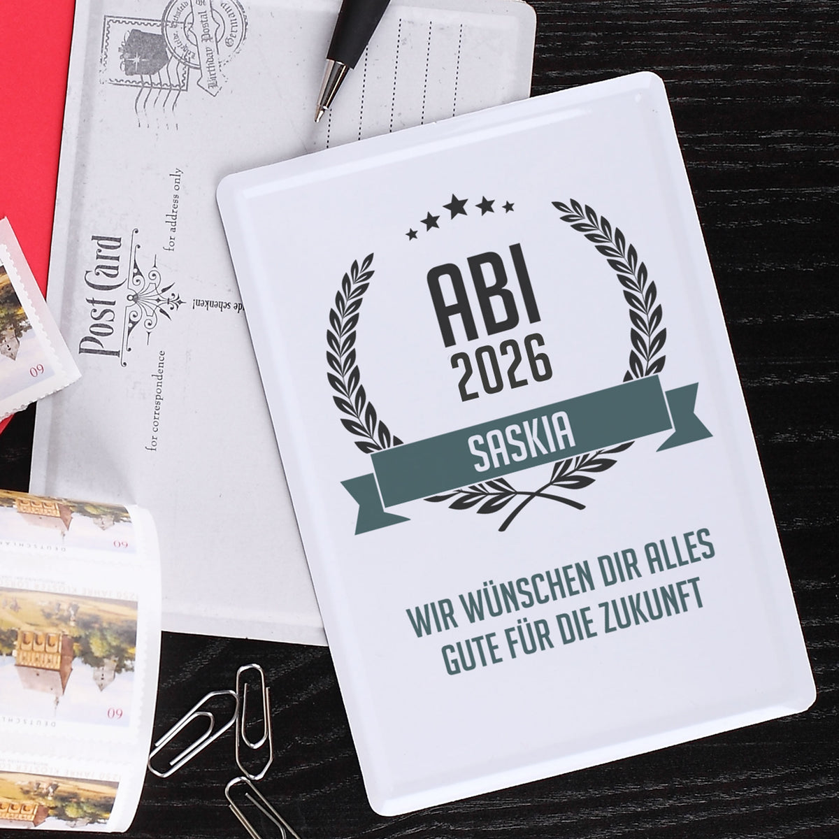 Abitur Blechpostkarte mit Glückwunschtext und Name auf dunklem Hintergrund mit Briefmarken und Büroklammern