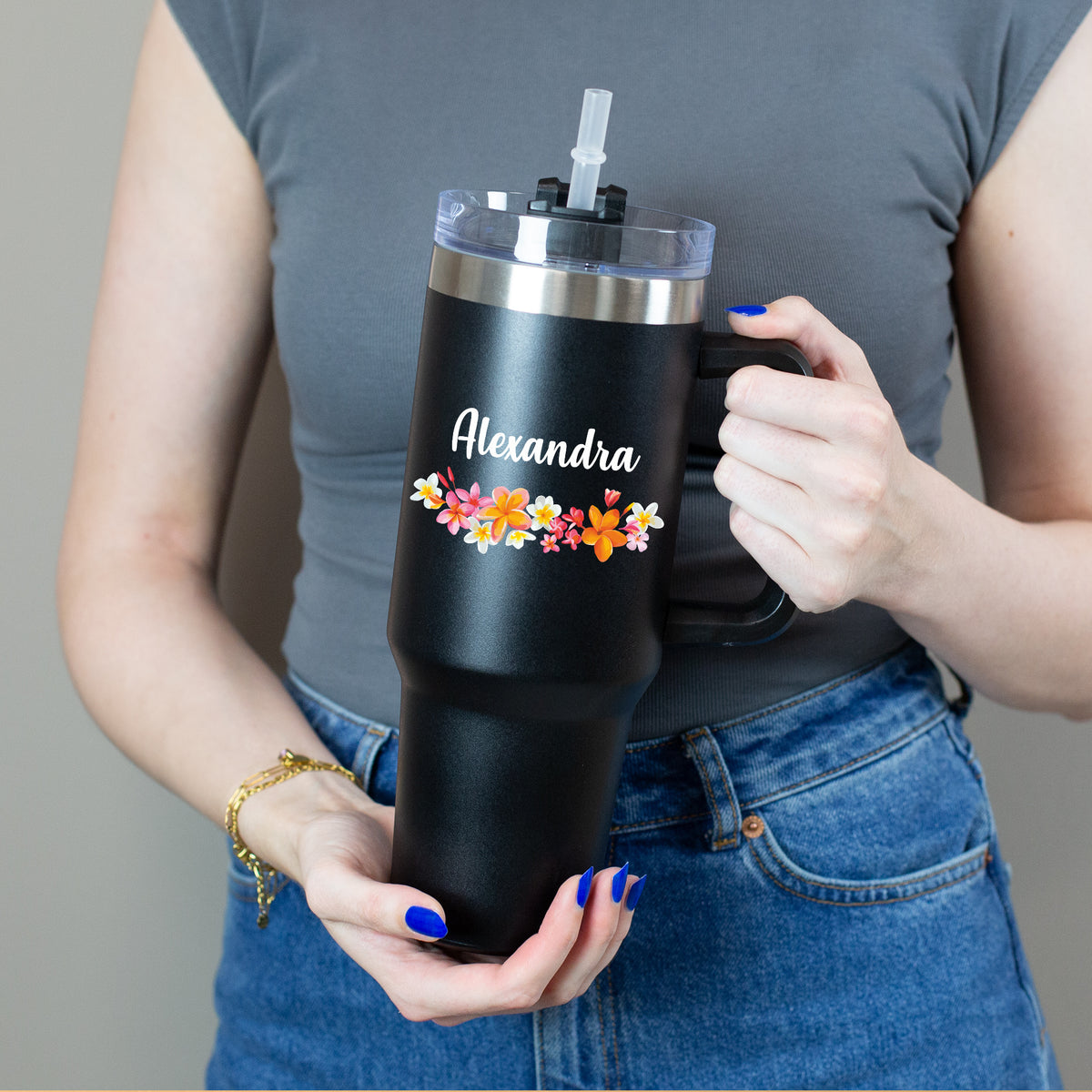 Person hält  Thermobecher mit buntem Blumenkranz und weißem Namen mit beiden Händen