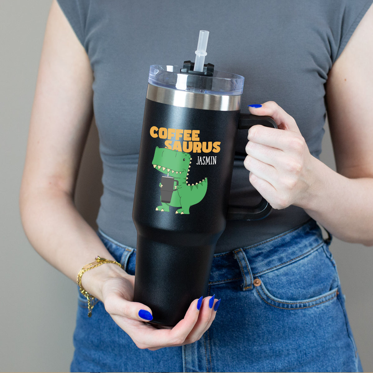 Person hält Thermobecher mit grünem Dino und „Coffee Saurus“-Schriftzug mit beiden Händen 