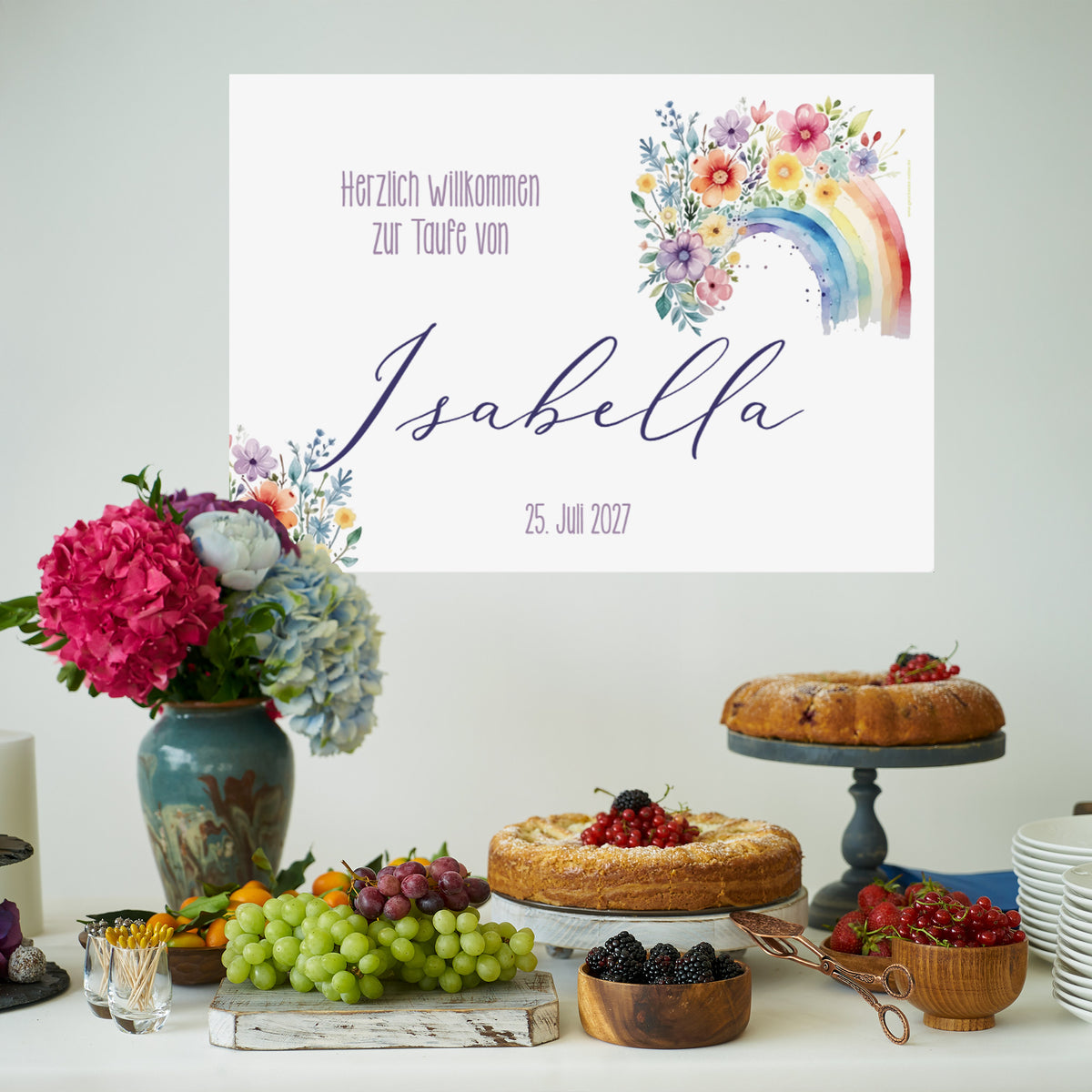 Dekoriertes Buffet mit Obst und Kuchen im Vordergrund, im Hintergrund hängt ein Banner mit Blumen-Regenbogenmotiv. Beschriftet mit „Herzlich willkommen zur Taufe von Isabella – 25. Juli 2027“.