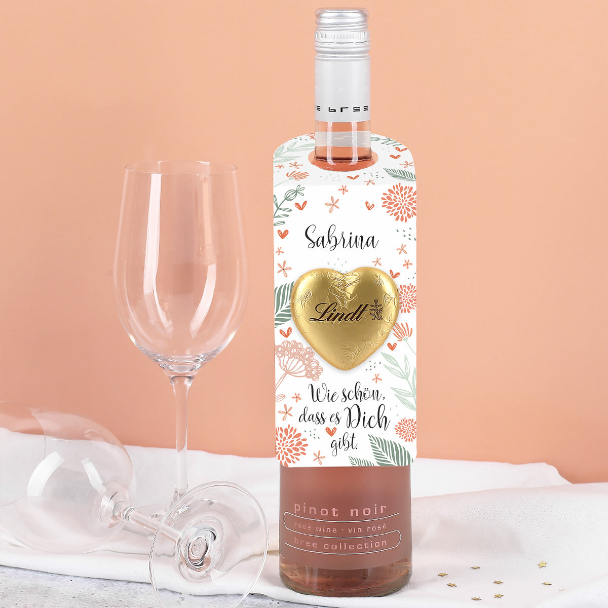 Flaschenanhänger im Blumendesign mit Lindt Schokoladenherz und dem Text „Wie schön, dass es dich gibt.“ – personalisiert mit Name, frontal an Flasche hängend.