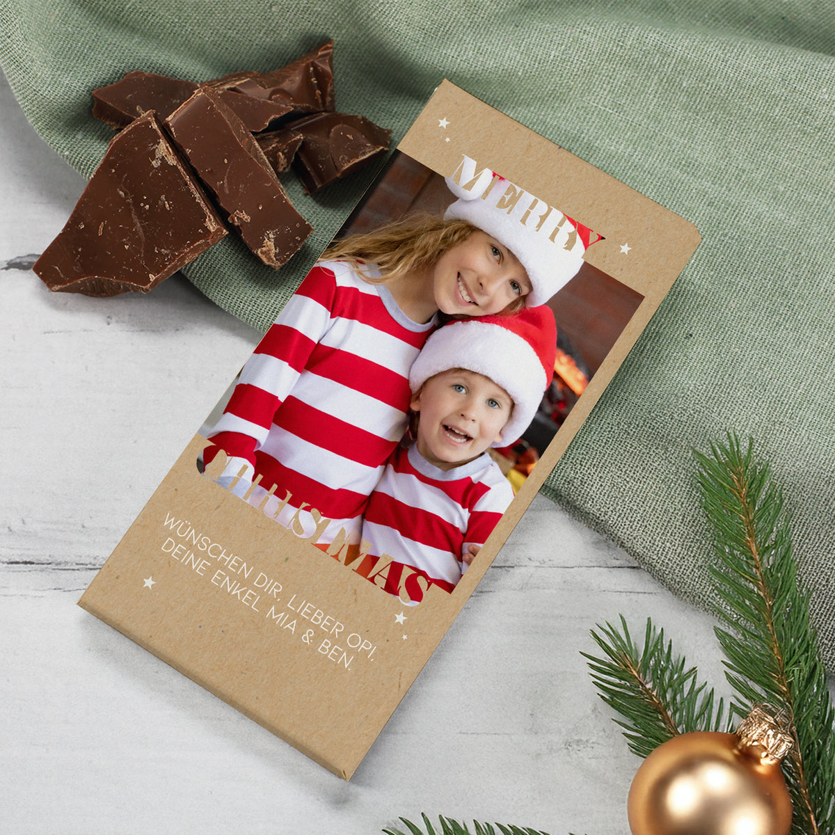 Personalisierte Schokoladentafel zu Weihnachten mit Foto und Wunschtext im „Merry Christmas“-Design. Die Verpackung besteht aus naturfarbenem Kraftpapier und ist mit weißen Sternen verziert. Auf dem Foto sind zwei Kinder mit Weihnachtsmützen abgebildet, daneben liegen Stücke feinster Vollmilchschokolade. Perfektes Geschenk zu Weihnachten, Nikolaus oder zum Wichteln.