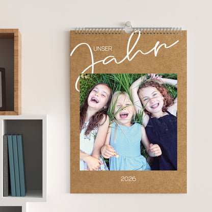 Foto-Wandkalender 2026 in Kraftpapier-Optik, aufgehängt an einer hellen Wand, mit Foto von drei lachenden Kindern und dem Titel „Unser Jahr“.