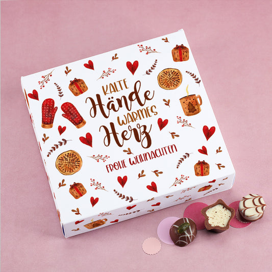 Personalisierbare Lindt Pralinenbox mit Aufdruck „Kalte Hände, warmes Herz – Frohe Weihnachten“, weihnachtlich dekoriert mit Herzen, Geschenken und Gebäck auf rosa Hintergrund.