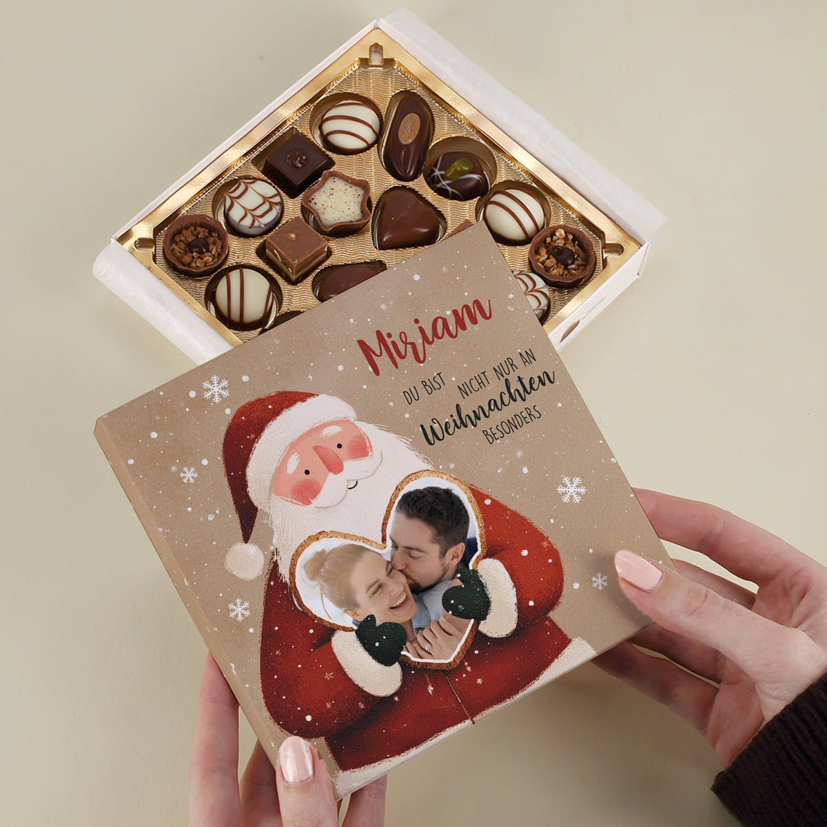 Weihnachtsmann hält ein Herz mit Foto eines lachenden Paares. Handschriftliche rote Schrift oben mit Name „Miriam“, daneben in Schwarz der Wunschtext. Der geöffnete Deckel zeigt fein sortierte Lindt Pralinen in goldener Pralinenschale.