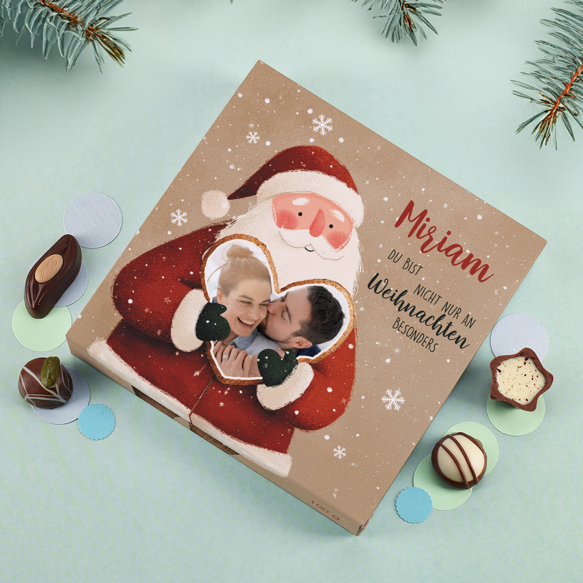 Weihnachtsmann mit Herzfoto eines Paares, umgeben von kleinen Schokoladenpralinen und runden Papierdekoren. Gut erkennbar: feine Schneeflocken im Hintergrund des Motivs.