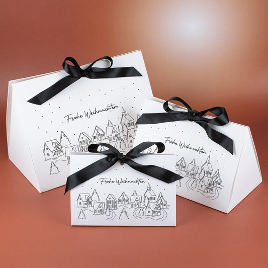 Drei Geschenktaschen in klein, mittel und groß mit Winterdorf-Motiv, Schneepunkten und schwarzer Satinschleife, vor warmem Hintergrund arrangiert.