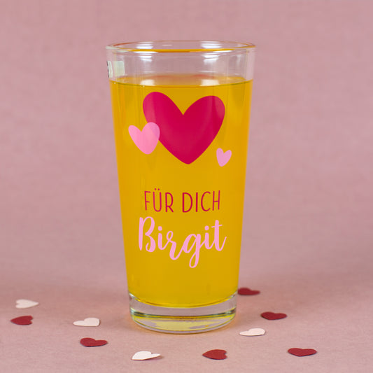 Personalisierbares Glas mit Herz-Motiv und Namen „Birgit“ auf rosa Hintergrund mit Dekoration