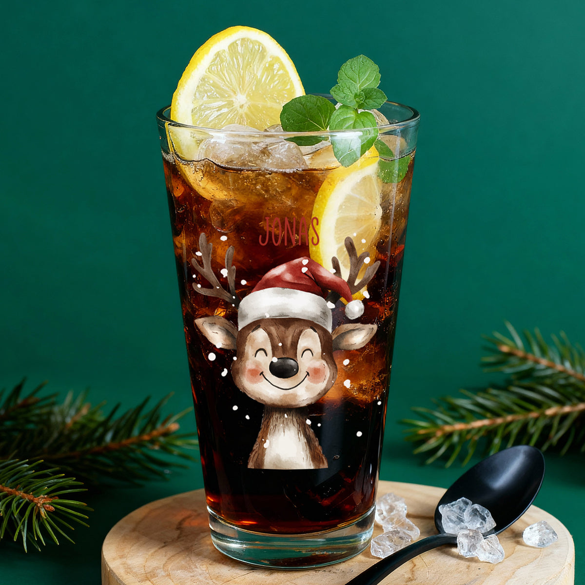 Weihnachtliches Trinkglas mit Rentier und Name Jonas, gefüllt mit Cola, Eiswürfeln und Zitronenscheiben vor grünem Hintergrund.