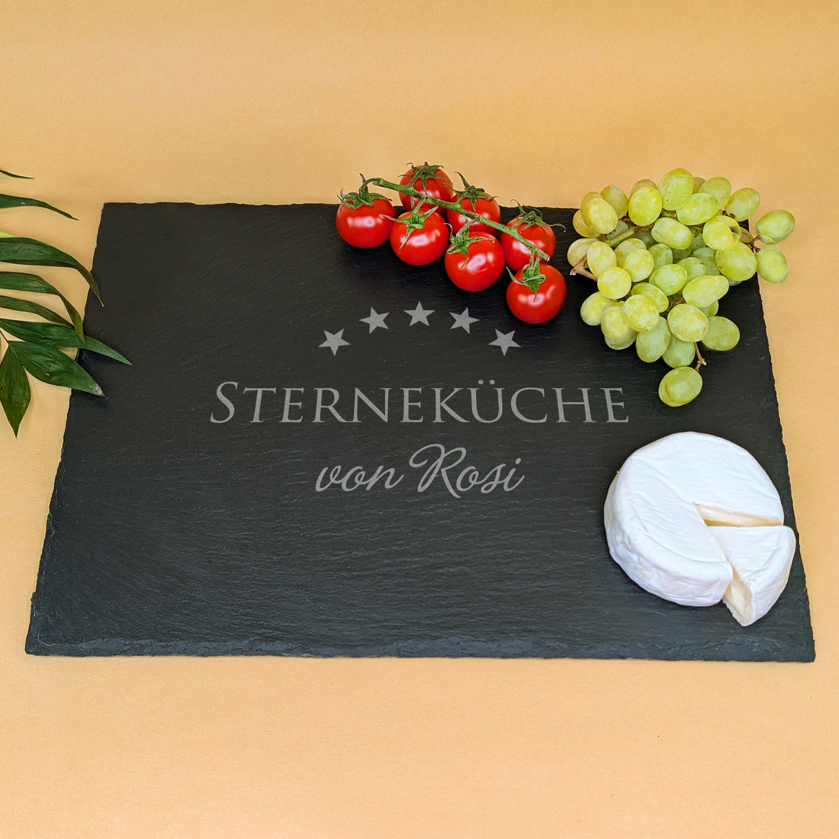 Schieferplatte mit Gravur „STERNEKÜCHE von Rosi“, dekoriert mit Tomaten, Trauben, Camembert und grünen Blättern – angerichtet auf beigem Hintergrund