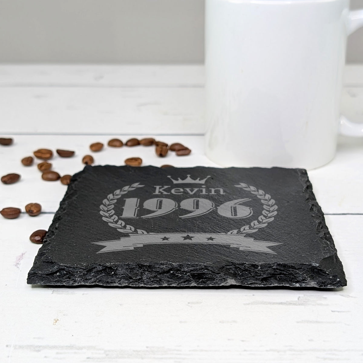 Schiefer-Untersetzer mit Gravur „Kevin 1996“ und Kranzmotiv, neben weißer Tasse und Kaffeebohnen