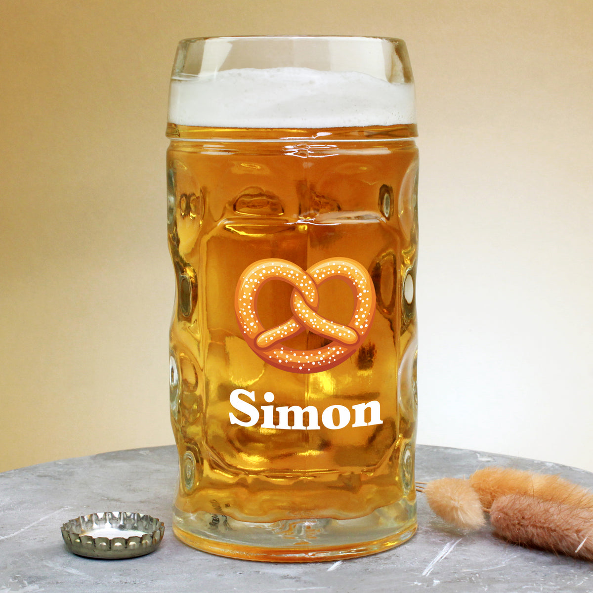 0,5-Liter-Bierglas mit Brezel-Motiv und Name Simon, auf Tisch mit Kronkorken und Dekozweig