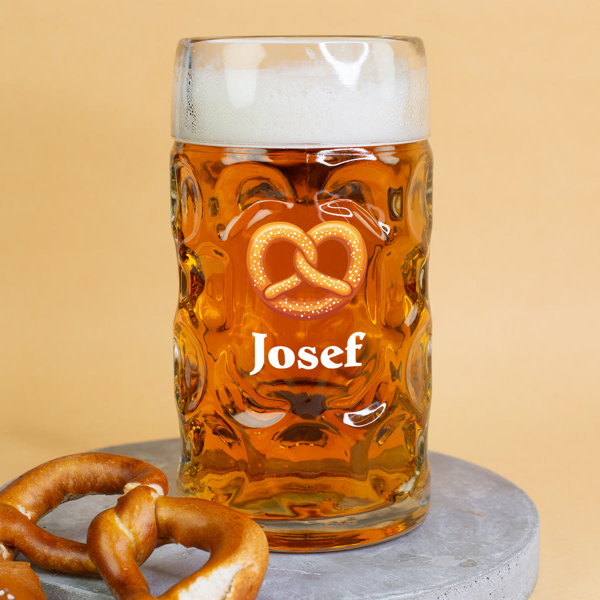 Großer Maßkrug mit Brezel-Motiv und Namensaufdruck Josef, schaumgekrönt auf runder Betonplatte mit Brezeln